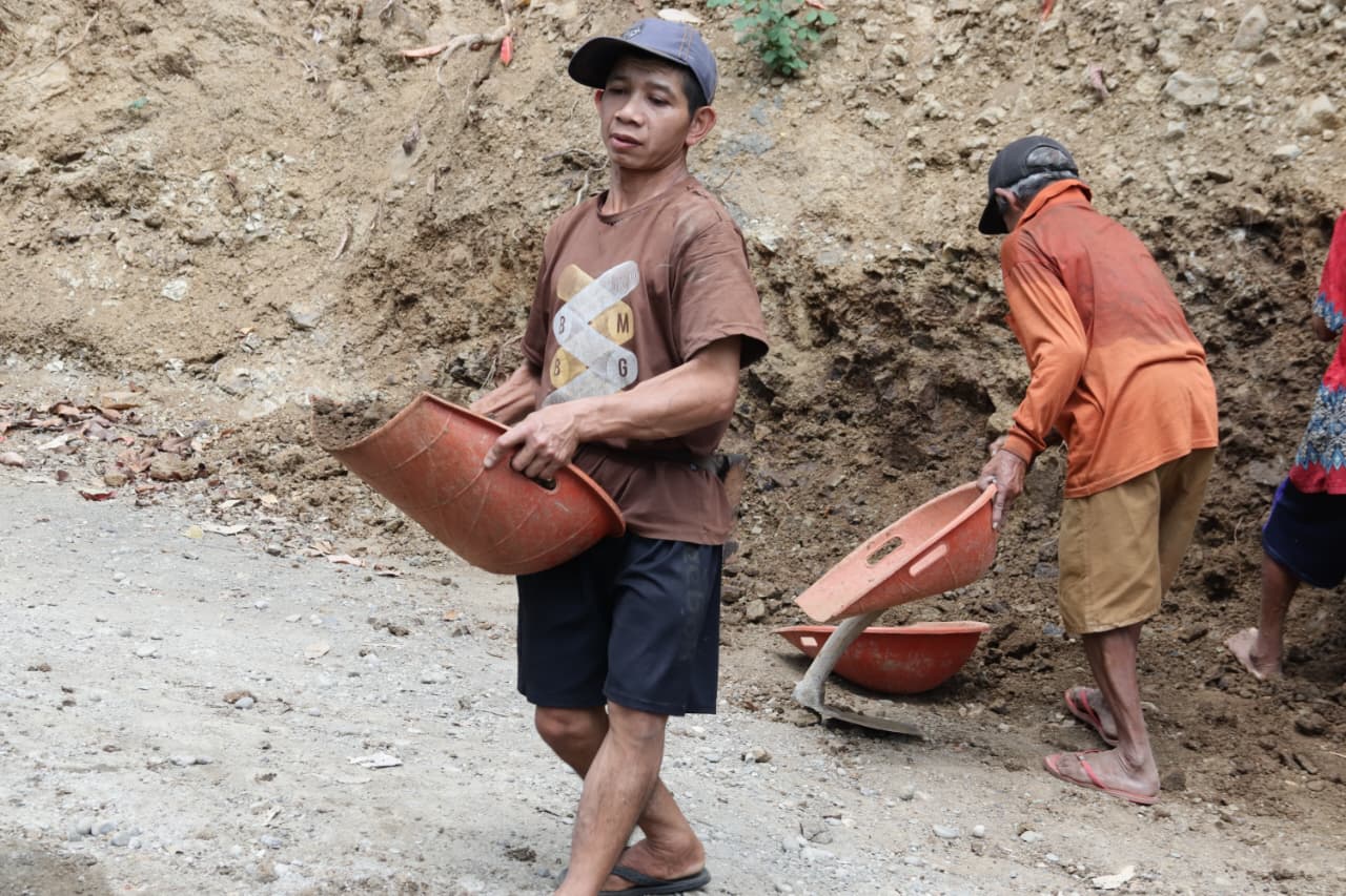 Minto tampak bolak-balik membawa tanah menggunakan alat sederhana menuju lokasi pekerjaan. Meski harus melewati jalur menanjak dengan beban yang cukup berat, semangatnya tidak surut. Baginya, setiap angkutan tanah merupakan kontribusi nyata bagi kemajuan desanya.