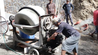 Madrohim, salah seorang warga, mengambil bagian dengan mengoperasikan mesin molen yang menjadi pusat pengolahan adukan beton, Minggu (26/7/2026).