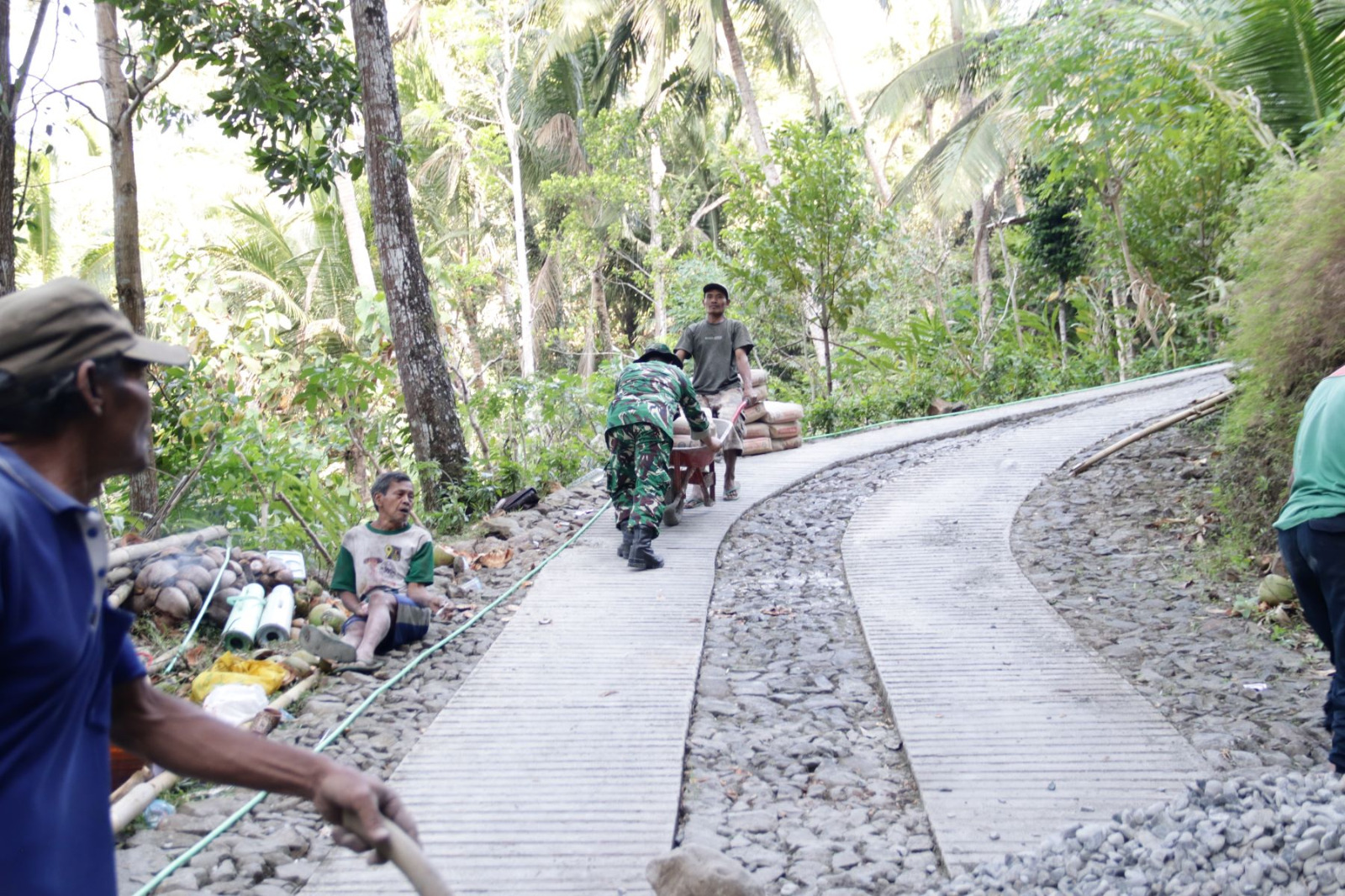 Desa Gumelem Kulon, Kecamatan Susukan, Serka Munasibun terlihat bahu-membahu bersama warga melangsir sak-sak semen menggunakan angkong menuju lokasi pengecoran jalan, Minggu (19/7/2026).