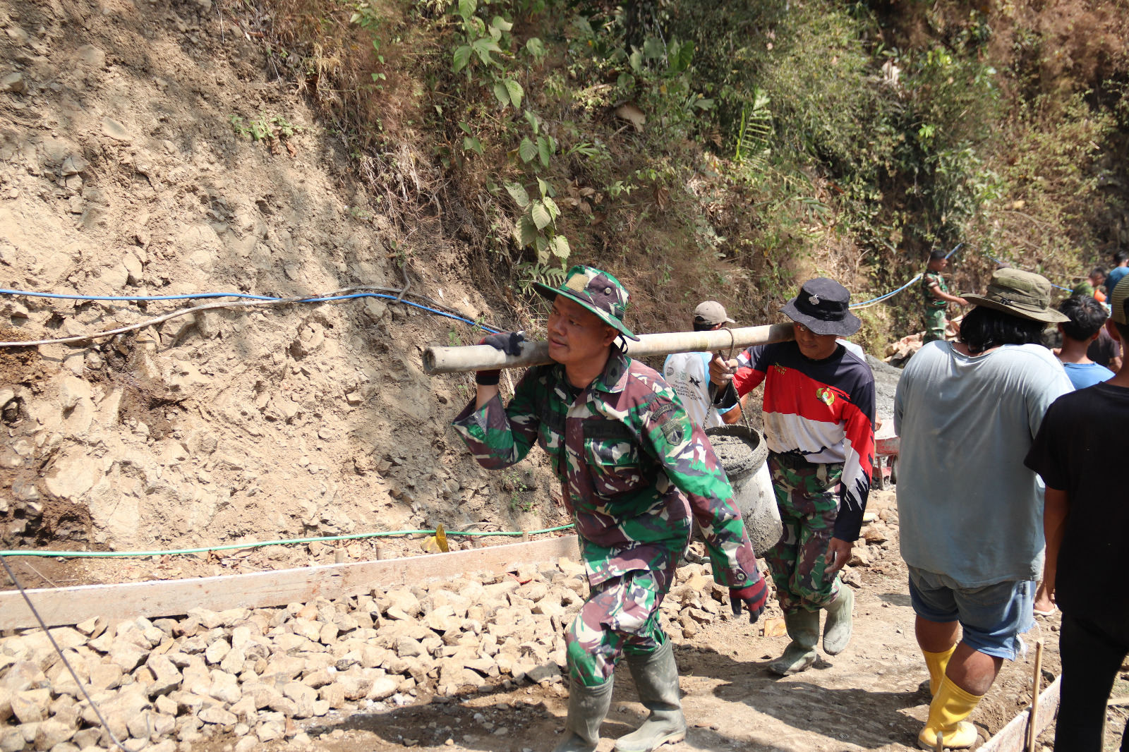 Totalitas Satgas TMMD: Pelda Ratam bersama rekannya memikul ember berisi adukan beton menuju lokasi pengecoran rabat beton di Dusun Kunir RT 01/09, Desa Gumelem Kulon, Kecamatan Susukan, Jumat (7/8/2026). Cara tersebut dilakukan untuk memperlancar distribusi material di medan yang sempit dan menanjak.